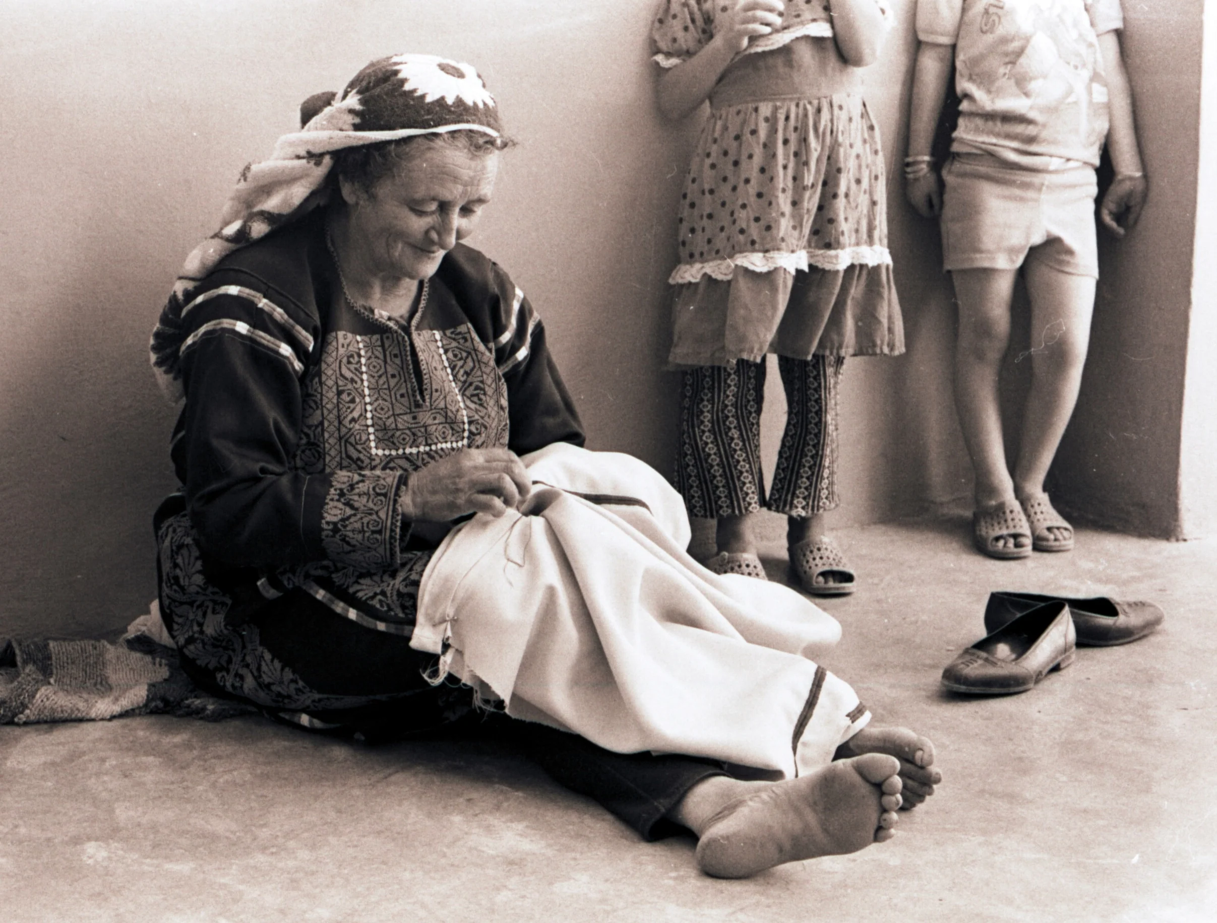 Fatima Yousef sewing a Palestinian thobe, seated on the floor in traditional dress, Kobar-Ramallah, 1970s